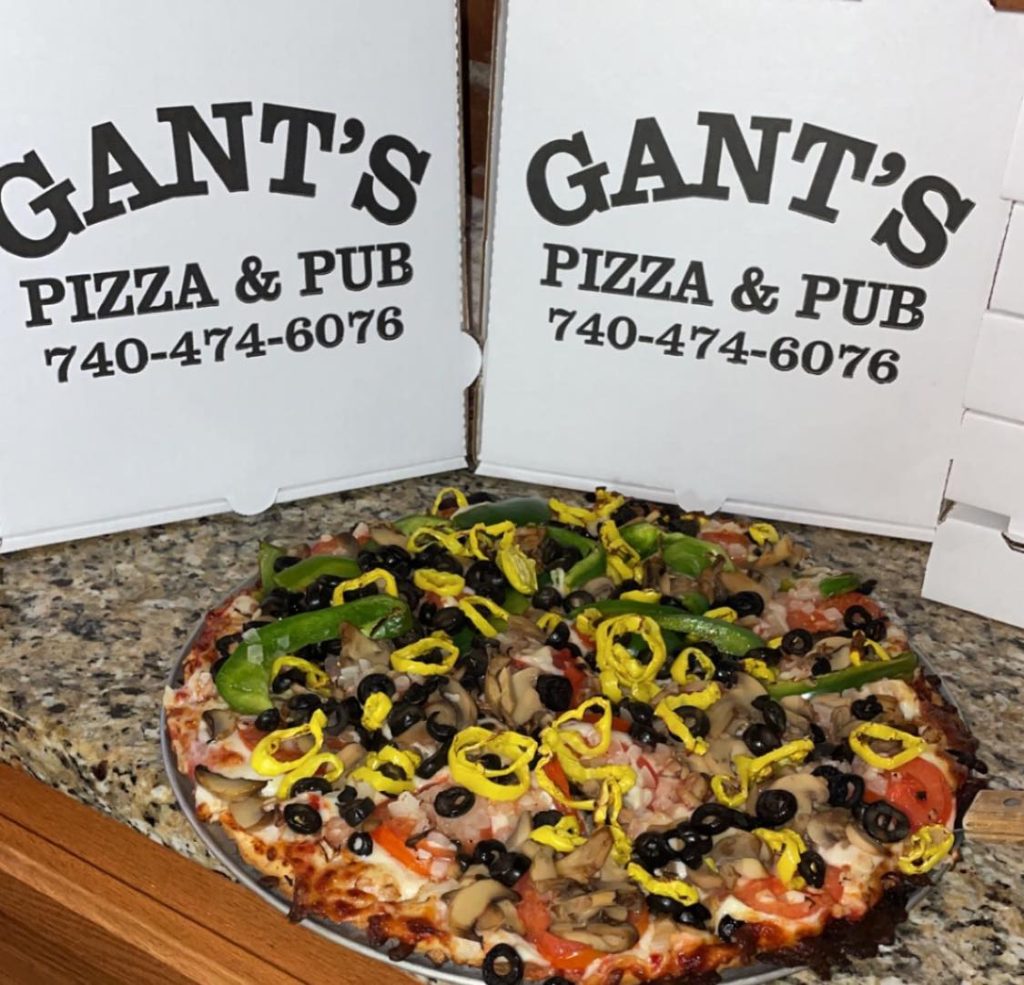 A photo of a loaded pizza with various toppings in front of two cardboard pizza boxes that say "Gant's Pizza & Pub" with their phone number on it: 740-474-6076.