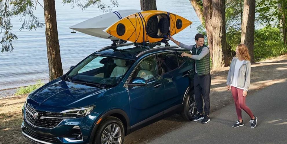 A blue 2022 Buick Encore GX with a canoe strapped on top. Two people, a man and a woman, are packing the car seemingly for a trip.