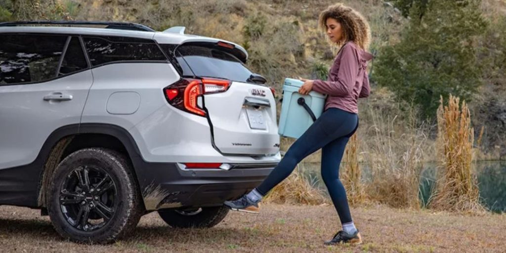 A woman in a pink shirt is using the hands-free trunk feature on the 2022 GMC Terrain to easily open it and store the box she is holding in her hands.