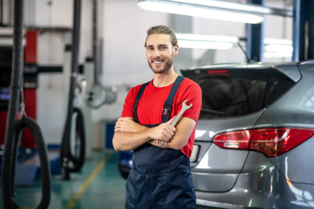 An auto repair technician holding a wrench standing in the auto repair shop of a dealership with a smile on his face.